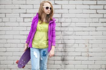 Blond teenage girl in jeans and sunglasses holds skateboard near gray urban brick wall