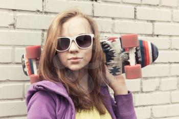 Blond teenage girl in sunglasses holds skateboard near gray urban brick wall, vintage tonal correction, old style photo filter effect