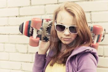 Blond teenage girl in sunglasses holds skateboard near gray urban brick wall, vintage tonal correction, old style filter effect