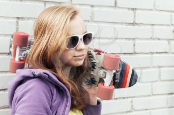Blond teenage girl in sunglasses holds skateboard near gray urban brick wall