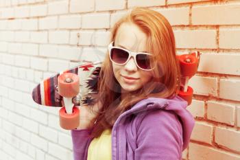 Blond teenage girl in sunglasses holds skateboard near gray urban brick wall, bright tonal correction, old style filter effect