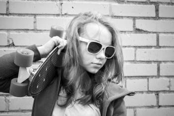 Blond teenage girl in jeans and sunglasses holds skateboard over urban brick wall, monochrome photo