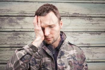 Young Caucasian man with headache in camouflage. Outdoor portrait over green rural wooden wall