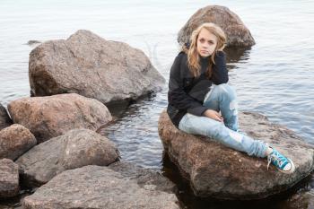 Beautiful blond Caucasian teenage girl sitting on coastal stones on the lake coast in Finland