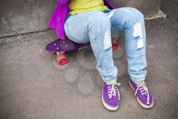 Teenager in blue jeans and gumshoes sits on skateboard