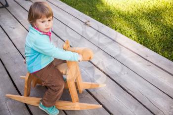 Outdoor portrait of cute Caucasian blond baby girl riding wooden horse toy