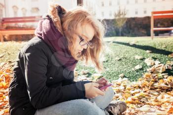 Cute Caucasian blond teenage girl in jeans, scarf and black jacket sitting in park and using smartphone for messaging, outdoor autumn portrait