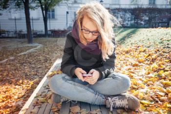 Cute Caucasian blond teenage girl in jeans and black jacket sitting on park bench and using smartphone, outdoor autumn portrait