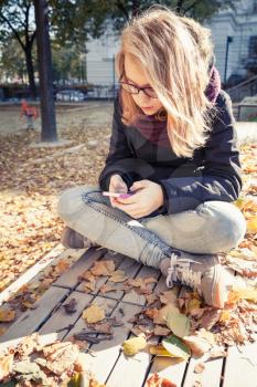 Cute Caucasian blond teenage girl in jeans and black jacket sitting on wooden park bench and using smartphone, outdoor autumn portrait
