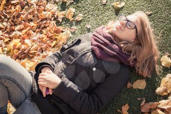Beautiful Caucasian blond teenage girl laying in autumnal park with falling leaves