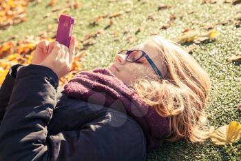 Caucasian blond teenage girl in black jacket laying in autumnal park and using cellphone for messaging in social network