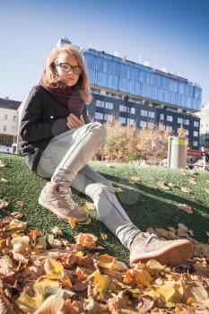 Beautiful Caucasian blond teenage girl in glasses sitting in autumnal city park with falling leaves