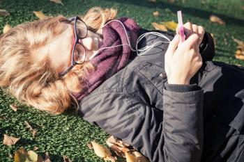Caucasian blond teenage girl in black jacket laying in autumnal park and using smart phone