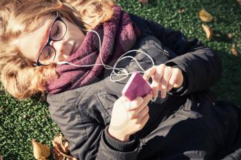 Caucasian blond teenage girl in black jacket laying in autumnal park and using smartphone