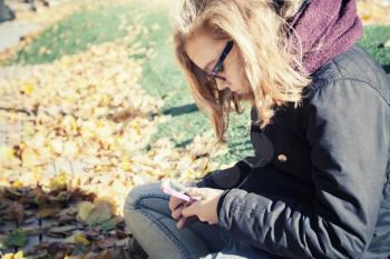 Caucasian blond teenage girl in glasses sitting in autumnal park and using smartphone, vintage style tonal correction photo filter