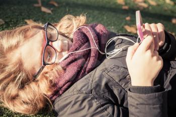 Caucasian blond teenage girl laying in autumnal park and using smart phone with headphones, vintage style tonal correction photo filter