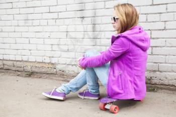 Beautiful blond teenage girl in sunglasses sits on skateboard near urban brick wall