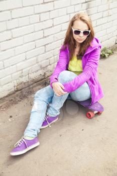 Beautiful teenage girl in sunglasses sits on skateboard near urban brick wall