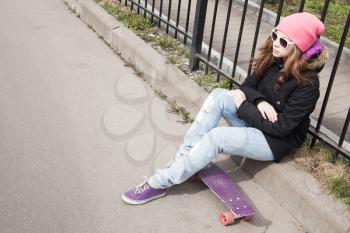 Teenage girl in jeans and sunglasses sits on her skateboard near urban fence