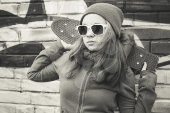 Teenage girl in sunglasses holds skateboard over urban brick wall background, monochrome photo