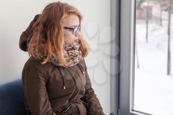 Beautiful blond Caucasian teenage girl in glasses and warm outwear sitting on blue sofa near winter window