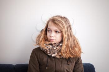 Beautiful blond Caucasian teenage girl in warm outwear sitting on blue sofa over white wall