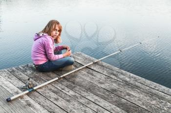 Blond Caucasian girl sitting on a wooden pier with fishing rod