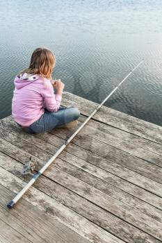 Little blond Caucasian girl sitting on a wooden pier with fishing rod