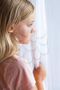 Close up vertical portrait of beautiful blond Caucasian girl standing near a window with white curtains