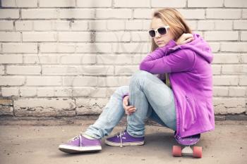Blond teenage girl in jeans and sunglasses sits on her skateboard near white brick wall, photo with warm retro tonal correction effect, old style filter