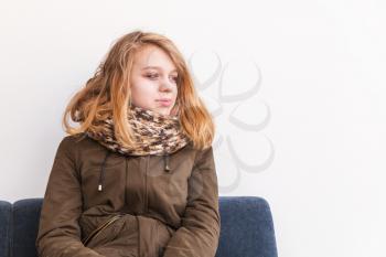 Beautiful blond Caucasian teenage girl in warm clothes sitting on sofa over white wall