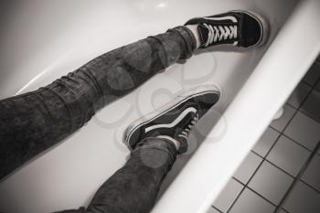 Teenager in black sporty sneakers lays in white bath, feet only. Black and white photo