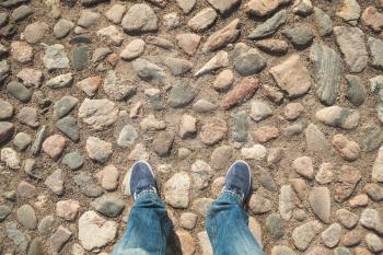 Male feet in blue jeans and sneakers on old rough stone pavement