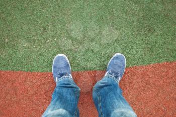 Male feet in blue jeans and sneakers standing on artificial stadium grass