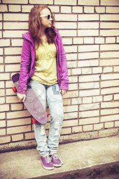 Blond teenage girl in sunglasses and colorful clothes with a skateboard near gray brick wall, vintage tonal correction photo filter, old style effect