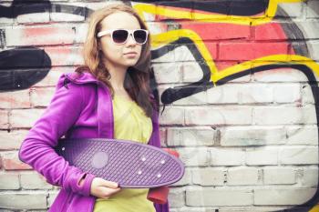 Blond teenage girl holds skateboard, portrait over urban brick wall