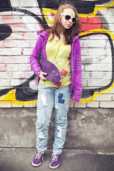 Blond teenage girl holds skateboard, vertical photo over urban brick wall