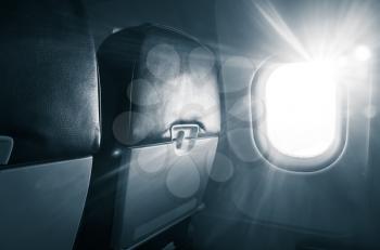Passenger plane interior fragment. Main cabin chairs with folding table and glowing porthole with natural sunlight lens flare. Blue toned photo