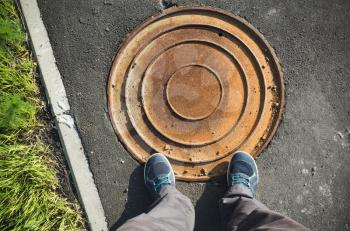 Male feet in blue sporty shoes stand on street pavement near round rusted sewer manhole cover