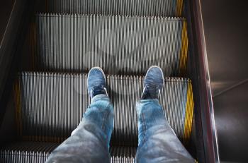 Male feet in blue jeans and sneakers stand on escalator stairs