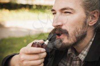 Young serious asian bearded man smoking pipe in summer park, close up portrait