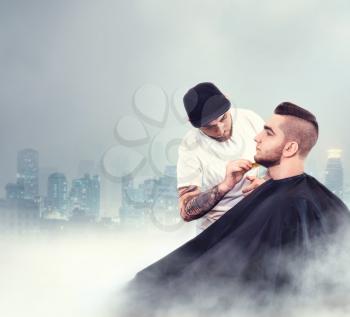 Young man having his beard shaven, barber working with comb over dark city background