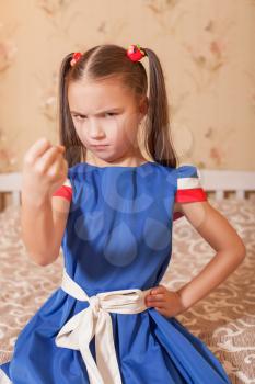 Angry little girl shows fist. Bedroom on the background.