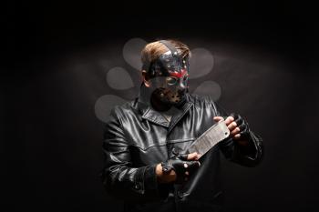 Portrait of bloody murderer in hockey mask holding a meat cleaver in hand against black background.