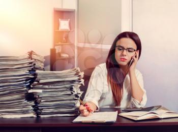 Young woman bookkeeper using mobile phone at workplace, stacks of documents on the table