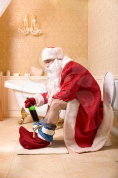 Funny drunk Father Christmas with bottle of alcohol sitting on the toilet.