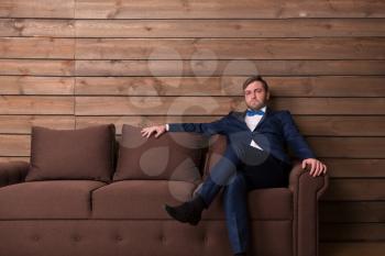 Serious groom in suit and bow-tie sitting on couch, wooden room interior on background