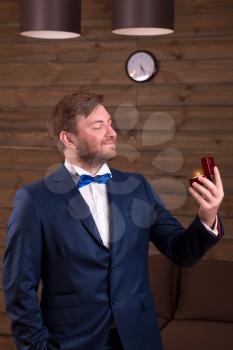 Happy groom in suit and bow-tie holding wedding ring in his hand, wooden room interior on background