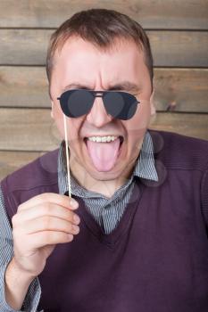 Playful man with funny sunglasses on a stick poses face, wooden background. Fun photo props and accessories for shoots
