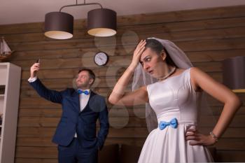 Dissatisfied bride and groom making selfie, wooden room on background.